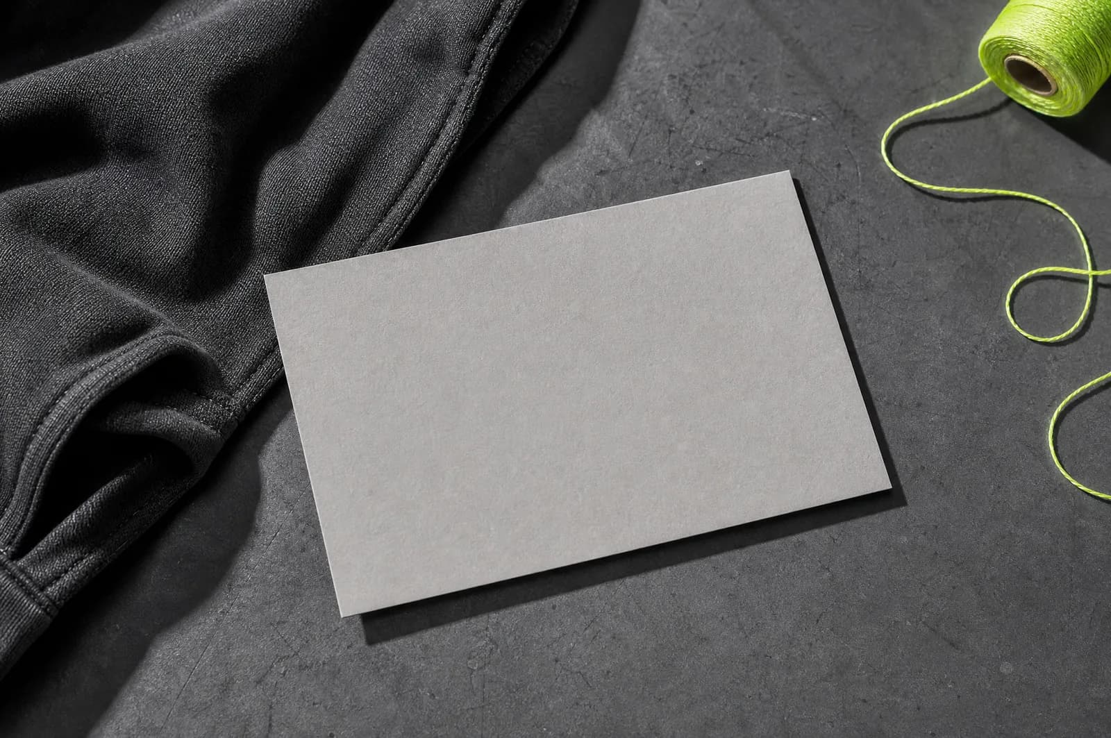A blank pale grey card lying on a graphite surface beside a folded garment edge and a hi-vis thread.